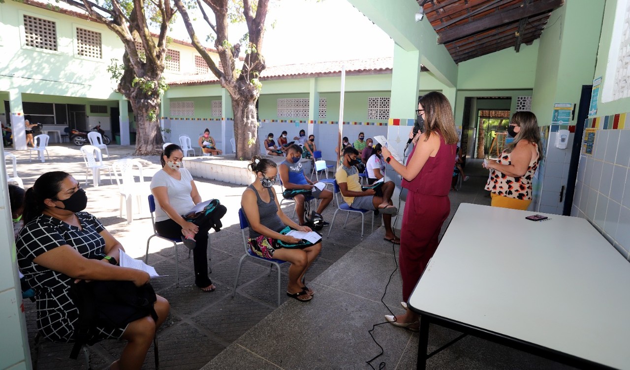 grupo de pessoas sentadas em cadeiras em um pátio de uma escola, as cadeiras estão distantes umas das outras e as pessoas usam máscaras